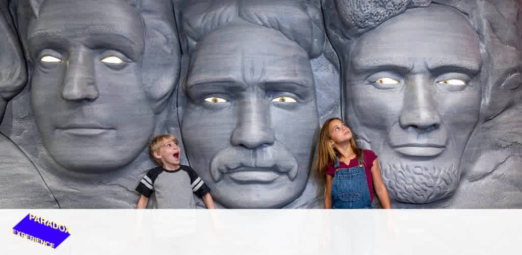 A young boy and girl stand in front of a wall featuring three large, sculpted faces with illuminated eyes. The boy is wide-eyed and open-mouthed, while the girl looks upward contemplatively. The sculpture exhibits detailed facial features creating a unique backdrop.