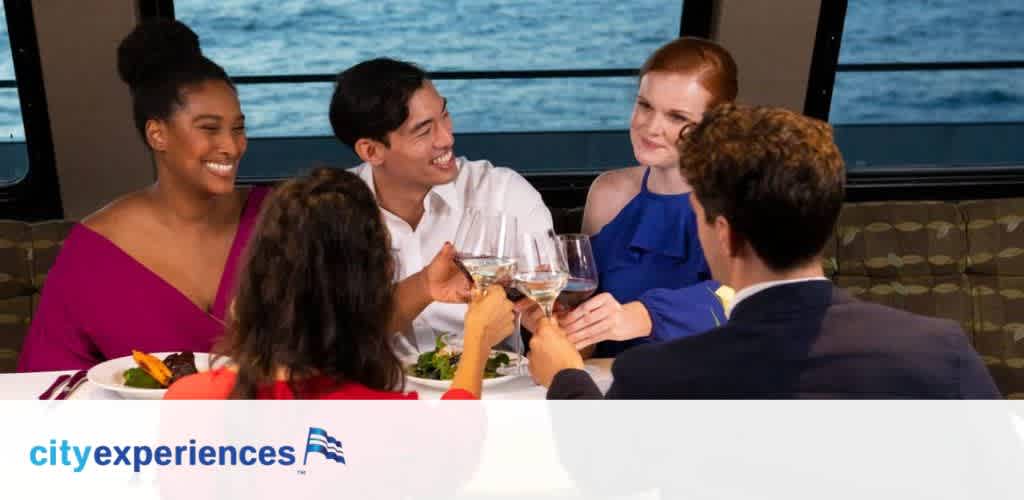 Image shows a group of four people enjoying a meal on a cruise. They are indoors, toasting with glasses, and smiling. There is a large window with a view of the water in the background. The  cityexperiences  logo with an icon of a flag is at the bottom center.