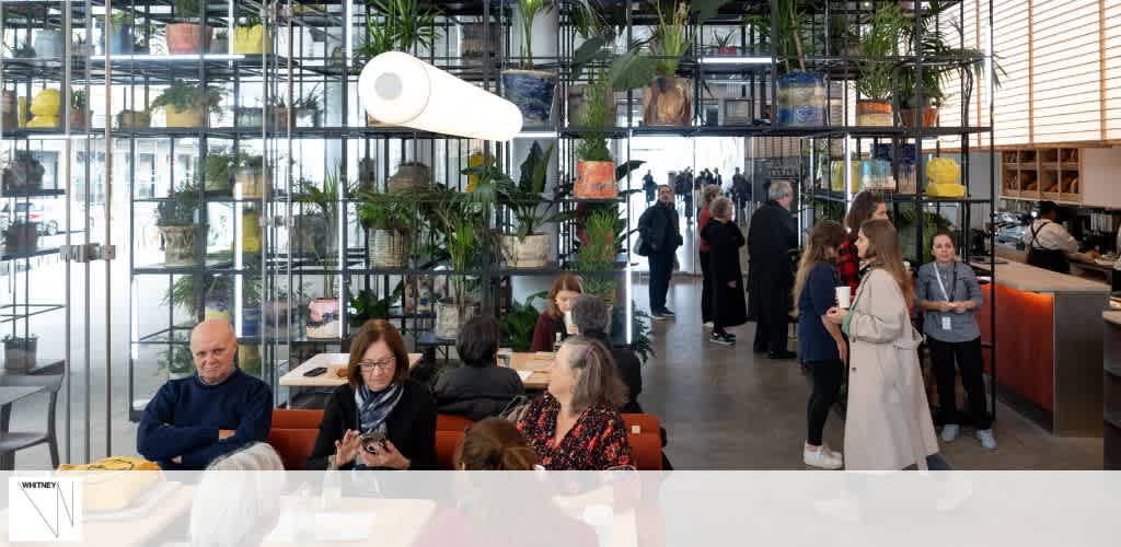 The image shows a casual indoor setting bustling with activity. Multiple groups of people are engaged in conversation while seated at tables with food and drinks. There is a prominent green touch due to various plants neatly organized on shelves that double as dividers, creating a cozy yet open atmosphere. A counter with staff attending to customers is visible in the background, adding to the dynamic and welcoming ambiance of the place.