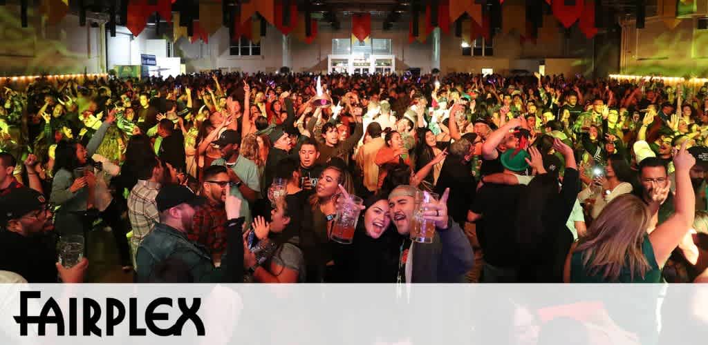 Image shows a lively crowd enjoying a nighttime event at Fairplex. Many attendees are raising their hands, waving to the camera, and surrounded by warm and green stage lights that create an inviting atmosphere. The bottom of the photo features the Fairplex logo.