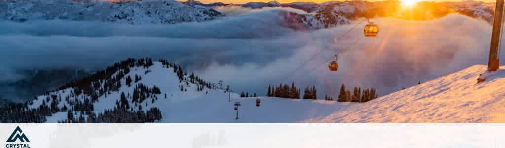 A snowy mountain landscape at sunset, featuring a ski lift with gondolas ascending. The sun casts a warm glow over the snow-covered slopes, with evergreen trees dotting the landscape. Clouds rest in the valley below, contrasting against the distant mountains in the background. The logo of a mountain with the word Crystal appears at the bottom left corner.