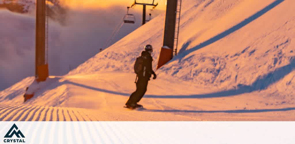A snowboarder descends a sunlit snowy slope near a ski lift. The warm, golden glow from the sun creates a captivating contrast with the crisp white snow. The slope shows distinct grooming lines, and the background features soft, misty clouds. A mountain logo is visible in the lower corner, enhancing the scene's adventurous spirit.