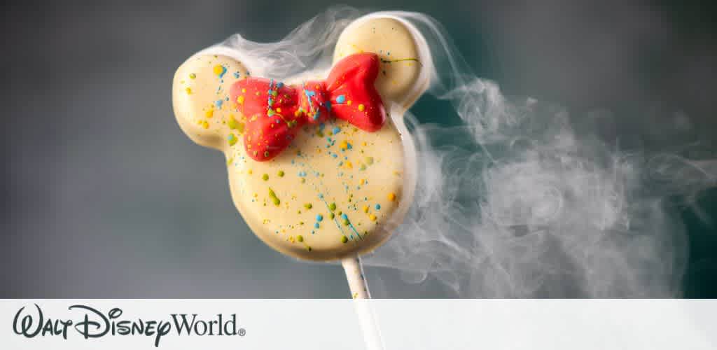 A whimsical dessert shaped like a familiar mouse head is creatively presented. Covered in white icing, it features colorful sprinkles and a red bow. Smoke or mist surrounds it, adding a magical effect. The Walt Disney World logo is displayed at the bottom, set against a neutral background.