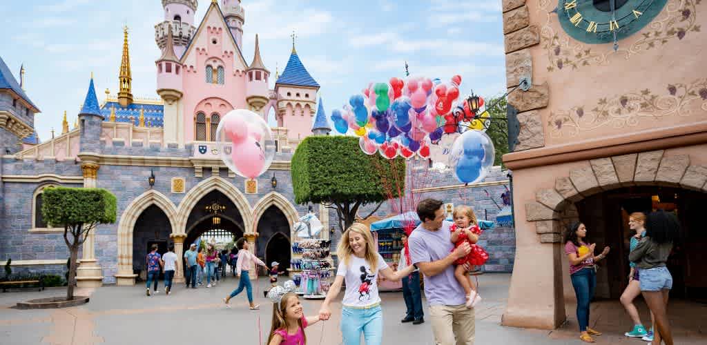 A joyful family strolls through a vibrant theme park, with a castle in the background. A woman holds a child's hand, while a man carries another child. Colorful balloons float nearby, and visitors explore the lively surroundings. Lush green trees and charming architecture enhance the enchanting atmosphere.