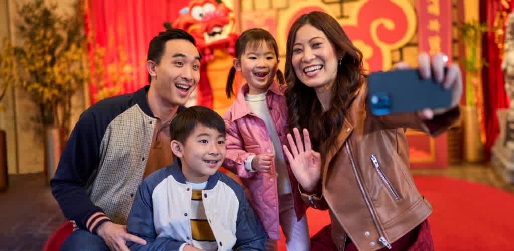 A cheerful family of four takes a selfie together indoors. They are smiling and appear to be enjoying a festive occasion. The background features red and gold decorations, including a dragon figure, suggesting a celebration. The family is casually dressed, adding to the relaxed and joyful atmosphere.