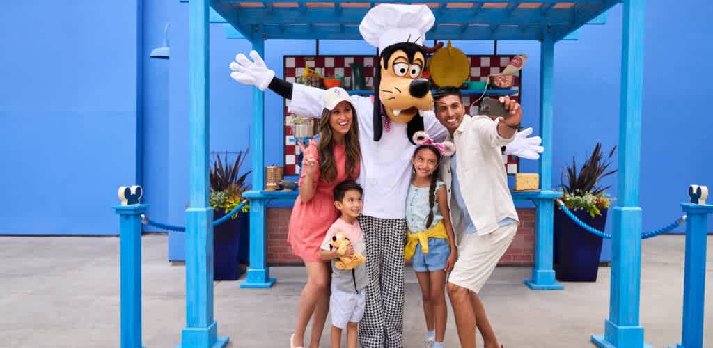 A family of four poses for a photo with a tall, cartoon character dressed as a chef. The setting is bright and colorful, with blue structures and decorative plants in the background. The family looks happy and excited as they smile and stand close together for the picture.