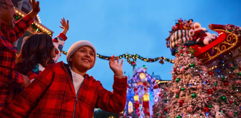 A joyful scene at a holiday parade, featuring a child in a red plaid jacket and white beanie waving enthusiastically. They are surrounded by festive decorations, including a brightly lit Christmas tree and a colorful float with a sleigh. The evening sky adds a warm backdrop to the celebration, enhancing the festive spirit.