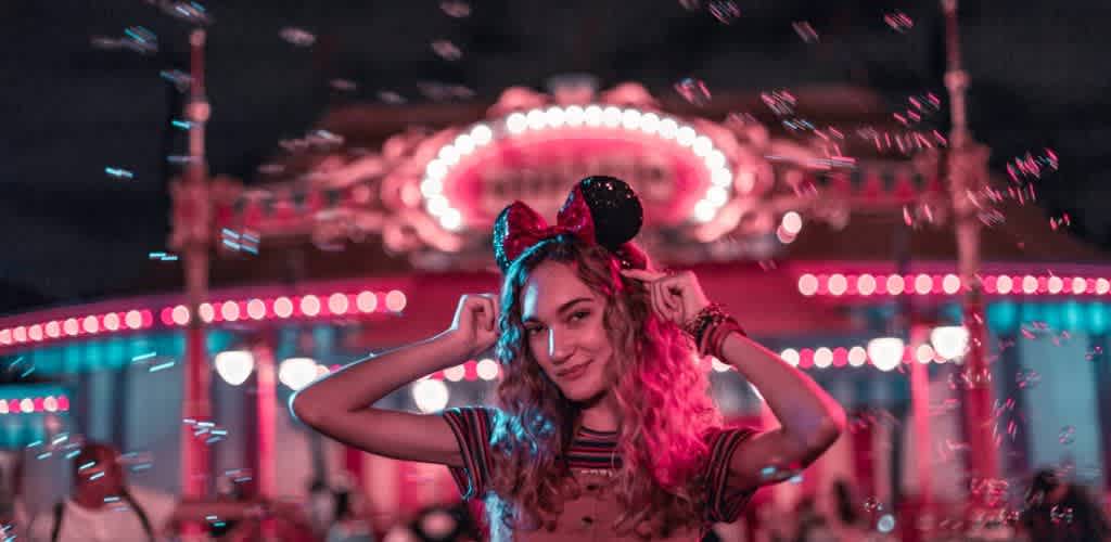 A young woman with long curly hair is wearing a headband with large mouse ears and smiling. She stands in front of a warmly lit amusement park carousel at night. Colorful lights illuminate the scene, and bubbles float in the air around her, adding a whimsical feel.