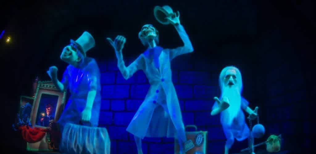 Three ghostly figures in bright blue glow stand against a dark brick background. The first figure wears a top hat and suit, holding a suitcase. The second figure wears a long coat, raising its hat. The third figure, with a long beard, gestures with one hand, holding a rounded object in the other. An ornate frame and red drapery are visible in the background on the left. The scene is vibrant and eerie, capturing a playful haunted theme.