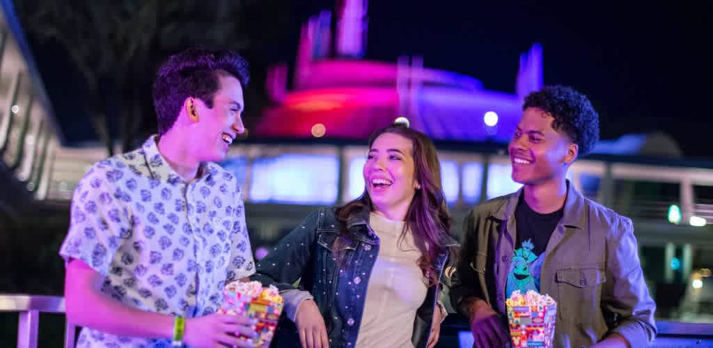 Three people enjoy a night out, smiling and holding popcorn. They stand outdoors with colorful lights illuminating the background, creating a fun and lively atmosphere.