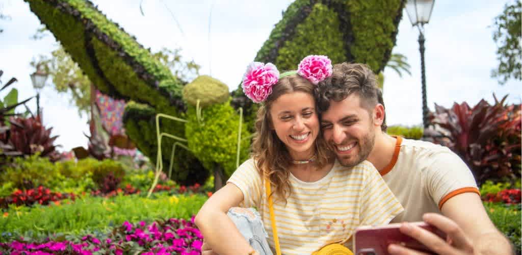 A couple smiles while taking a selfie in a vibrant garden setting. The woman wears pink floral headbands, and both are dressed casually. Behind them is a large artistic topiary shaped like an insect, surrounded by colorful flowers. The atmosphere is joyful and relaxed, suggesting a fun day outdoors.