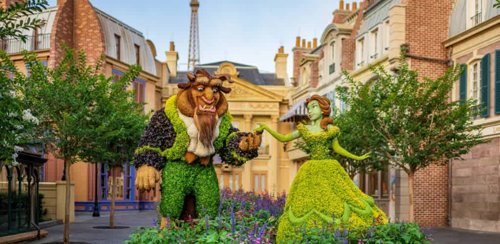 A lush topiary display portrays a graceful woman in a green dress and a large, beast-like figure holding hands, set amid colorful flowers. The scene is framed by charming European-style buildings in a quaint street setting, with the Eiffel Tower in the background. The sky is clear, adding to the vibrant and whimsical atmosphere. Perfect for a magical outdoor park or garden theme.