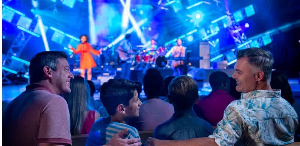 A lively concert scene with a diverse audience enjoying a performance. In the foreground, two men and a boy are smiling, engaged in the event. The stage is vibrant with colorful lights and musicians performing energetically. The setting is dynamic, filled with blue and purple hues that add excitement to the atmosphere. The image captures the essence of fun and togetherness in a live music experience.