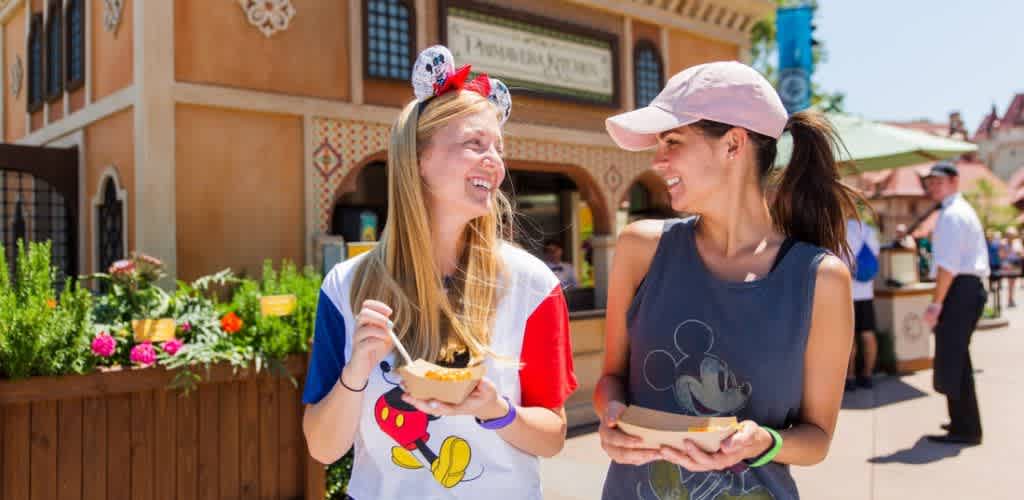 Two people smiling and enjoying food at an amusement park. One is wearing a colorful T-shirt with cartoon graphics and headband, while the other is in a sleeveless top and baseball cap. They stand near a beautifully designed building surrounded by flowers, exuding a joyful and relaxed atmosphere under clear blue skies.