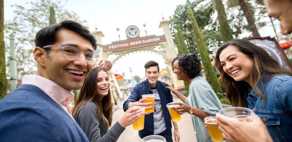 A group of six people stand outdoors enjoying a lively gathering at the Festival Beer Garden. They are smiling and raising plastic cups filled with beer, suggesting a celebratory moment. The background features an inviting arch with trees, enhancing the festive atmosphere. The people appear joyful and engaged with each other, capturing the essence of a fun social experience.