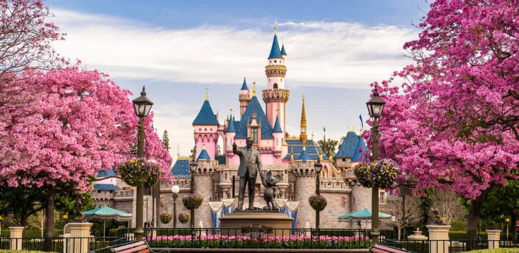 The image shows a whimsical castle with pink spires and blue rooftops surrounded by bright pink flowering trees. A statue stands in front of the castle amid a landscaped garden. The setting is vibrant and enchanting, evoking a fairytale atmosphere against a blue sky backdrop.