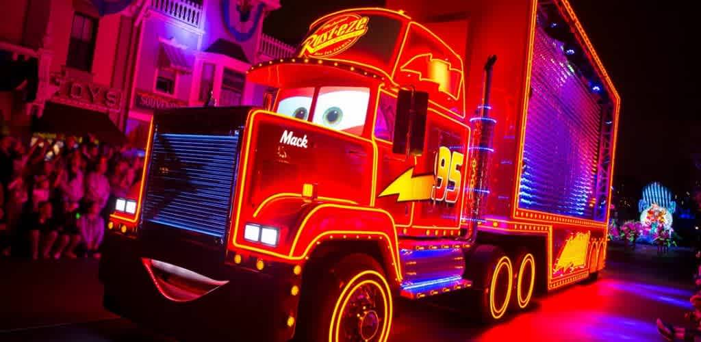 A vibrant, illuminated red truck designed to look like a character from a popular animated film drives through a lively night parade. The truck features colorful neon lights outlining its shape, visible smiling face, and animated eyes on the windshield. Crowds of people are seen in the background, enjoying the festive atmosphere, with buildings and other parade elements bathed in colorful lights.