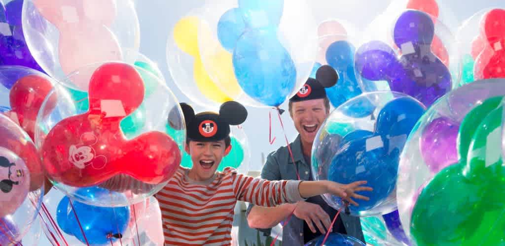A joyful scene featuring two people wearing mouse-ear hats surrounded by an array of colorful balloons. The balloons are shaped like mouse heads and come in vivid colors such as red, blue, purple, and green. The individuals appear excited and happy, with one of them reaching out towards the camera. Bright sunlight enhances the cheerful atmosphere.