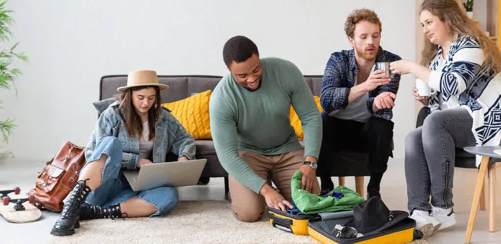 A group of four friends are in a cozy living room. One person sits on the floor packing a suitcase, while another looks at a laptop. Two others are seated on a sofa, one offering a drink to the other. A skateboard and backpack are on the floor. The setting is bright and casual, suggesting preparation for a trip.