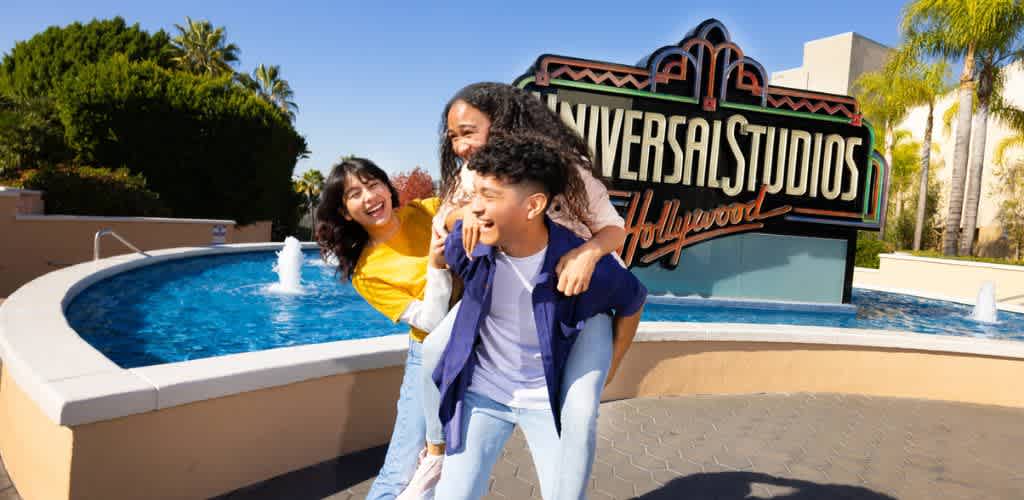 A group of three friends enjoys a sunny day at Universal Studios Hollywood. One person gives a playful piggyback ride to another, while all three share a laugh. In the background, a large Universal Studios Hollywood sign stands next to a blue fountain. Surrounded by lush greenery and palm trees, the scene is lively and fun, evoking a sense of adventure and excitement.