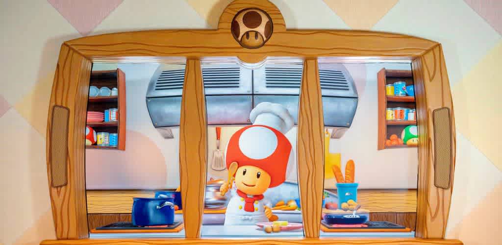 A cheerful cartoon character with a red mushroom cap stands in a colorful kitchen. The kitchen is equipped with a stove, blue pots, and various kitchen items. Shelves on both sides hold canned goods and vegetables. The kitchen window features a brown wooden frame with a mushroom emblem at the top, adding a whimsical touch. The scene is bright and engaging, perfect for a family-friendly setting.