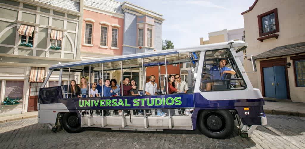 A Universal Studios tour tram is pictured with a group of diverse people sitting inside, enjoying the ride. The tram is parked on a cobbled street surrounded by colorful, whimsical buildings with decorative awnings and windows. The scene conveys a lively and inviting atmosphere, perfect for exploring and enjoying the attractions at Universal Studios.