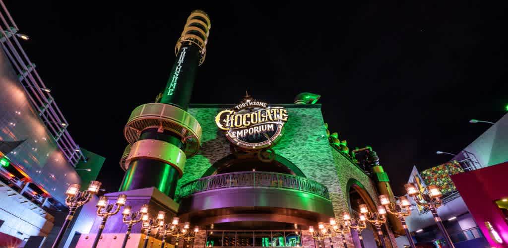 The image showcases an illuminated building at night, featuring a large sign that reads Chocolate Emporium. The architecture is whimsical with industrial elements, including a tall smokestack and glowing lights. A row of vintage-style street lamps lines the entrance, enhancing the enchanting ambiance. The night sky provides a dark backdrop, making the building's vibrant colors stand out.