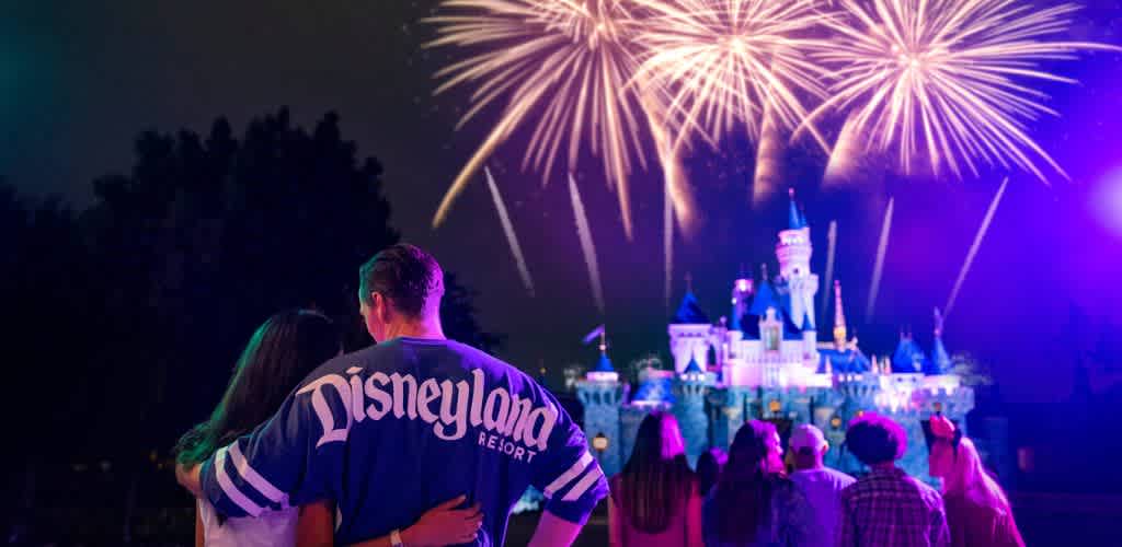 A couple stands with their backs to the camera, embracing as they watch a fireworks display over a brightly lit castle at night. The castle is iconic and surrounded by other park visitors, creating a lively atmosphere. The sky is illuminated by the colorful fireworks, adding a magical touch to the scene.