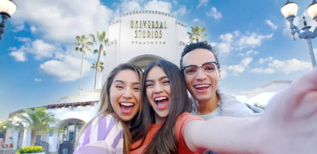 Three friends are smiling and taking a selfie at the entrance of Universal Studios Hollywood. The iconic archway and palm trees are visible in the background under a clear blue sky with a few scattered clouds. The group appears joyful and excited, capturing a fun moment at the theme park.