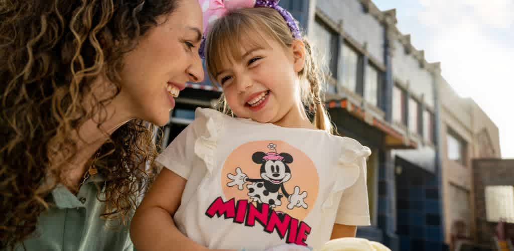 A joyful mother and daughter share a happy moment outdoors. The mother with curly hair smiles at her daughter who is wearing a white shirt with a cartoon character and pink bow, laughing with her eyes closed. Behind them are buildings with blue sky visible. The image conveys warmth and a sense of togetherness, perfect for showcasing family-friendly experiences.