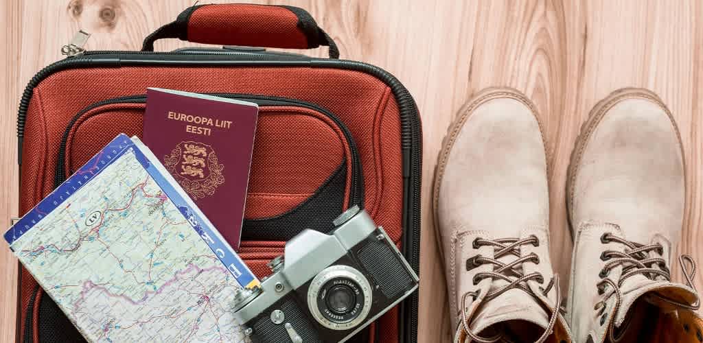 A pair of tan hiking boots is placed next to a red suitcase on a wooden floor. On the suitcase, there is a map, a passport, and a vintage camera, suggesting travel and adventure. The image conveys readiness for a trip, with essential travel items arranged neatly.