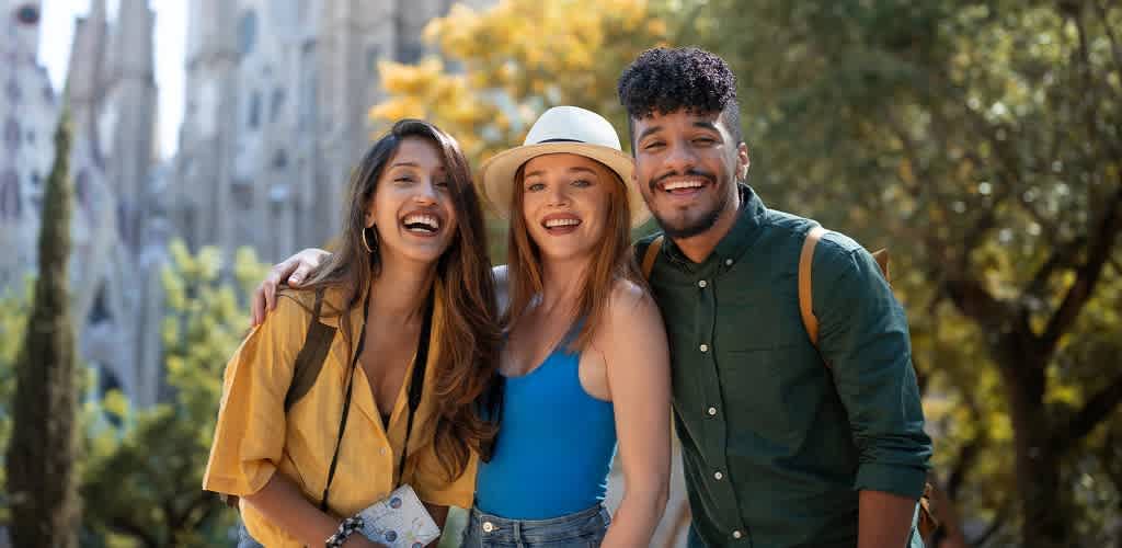 Three friends are standing together outdoors, smiling brightly. One wears a yellow shirt, another a blue top with a hat, and the third a green shirt. They are surrounded by lush greenery and sunlight, creating a cheerful and vibrant atmosphere, perfect for a fun outing on a sunny day.