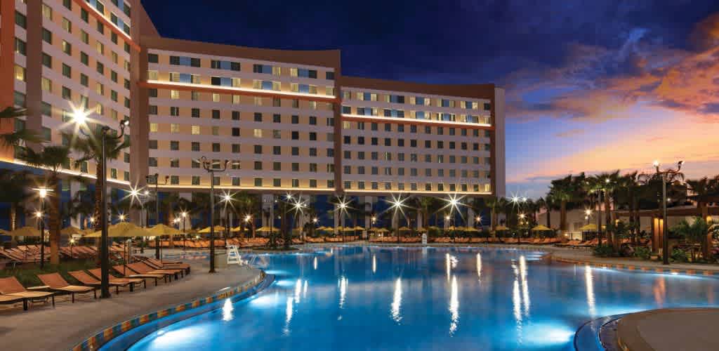 A twilight view of a large hotel building with numerous lit windows, standing adjacent to a vast, illuminated swimming pool. The pool is surrounded by loungers and palm trees, creating a serene and inviting atmosphere. The sky is a mix of deep blue and warm orange hues, enhancing the tranquil setting.