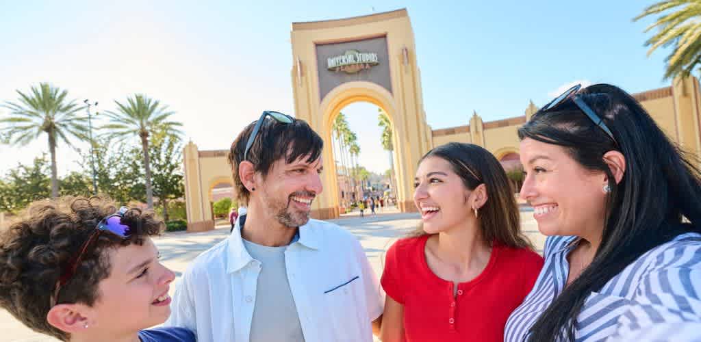 A family of four happily stands in front of a theme park entrance, surrounded by palm trees under a clear blue sky. The two adults and two children are smiling and looking at each other, conveying excitement and joy. The park's large archway features signage in the background, adding to the lively atmosphere of a fun family outing. The bright, sunny day enhances the cheerful scene.