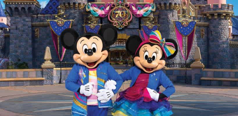 A joyful image featuring two iconic cartoon mice in colorful outfits standing in front of a castle adorned with decorative banners. The characters are smiling, with one wearing a rainbow hat and the other in a suit. The scene conveys a festive atmosphere, ideal for an amusement park or celebration setting.