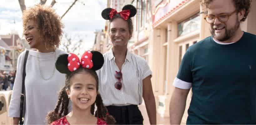 A group of four people are joyfully walking down a lively street, including a girl wearing Minnie Mouse ears with a red bow. The woman next to her and another woman in the group are also wearing Minnie Mouse ears. The atmosphere is bright and cheerful, with colorful buildings and a sunny sky in the background, creating a fun and festive environment perfect for an amusement park visit.