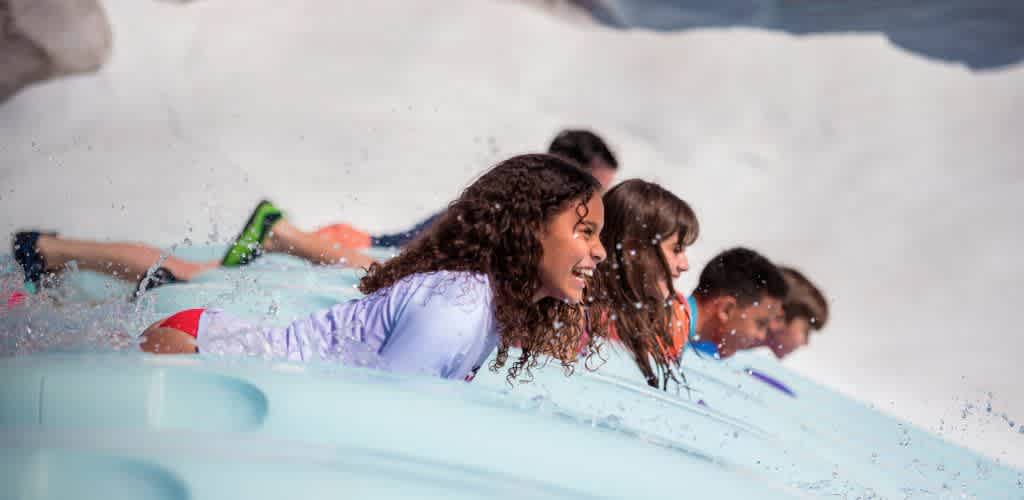 A group of people enjoy a thrilling ride down a water slide. They are lying on blue mats, racing side by side, with smiles and excitement on their faces. Water splashes around them as they descend, creating a lively and fun atmosphere. The background is a blur, capturing the speed and exhilaration of the activity. Perfect for showcasing the excitement at Funex water park.