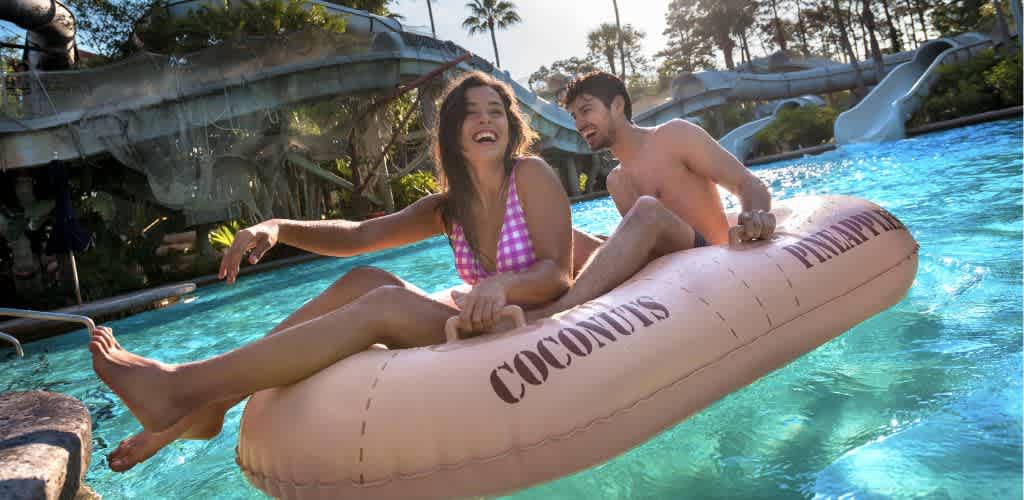A woman in a pink swimsuit and a man in swim trunks enjoy a summer day at a water park, riding a large inflatable tube in a pool. The sun is shining, creating a lively atmosphere with slides and greenery in the background. The woman smiles broadly while the man sits behind her, both appearing joyful and relaxed. The pool water glistens in the sunlight, emphasizing the fun and excitement of the experience at the park.