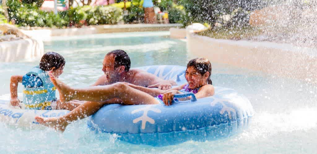 A family enjoys a sunny day at a water park, floating on a lazy river. Two children and an adult are on a large blue inflatable tube with playful designs. Lush greenery and sprays of water surround them, creating a refreshing and joyful atmosphere. The scene captures the essence of summer fun and leisure.