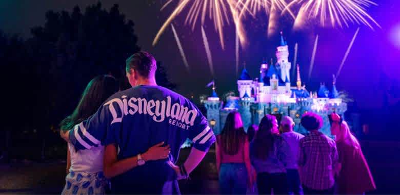 A group of people stands in front of a brightly lit castle at night, watching a vibrant fireworks display light up the sky. The colorful fireworks create a festive atmosphere. The group, including a person wearing a jersey with text on the back, is enjoying the spectacle while embracing and taking in the magical scene.