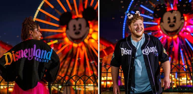 Two people stand in front of a brightly lit Ferris wheel featuring a large Mickey Mouse face. The person on the left faces away wearing a colorful Disneyland Resort jacket, while the person on the right smiles, wearing mouse ears and a Disneyland jacket over a light blue shirt. The background is vibrant with colorful lights.