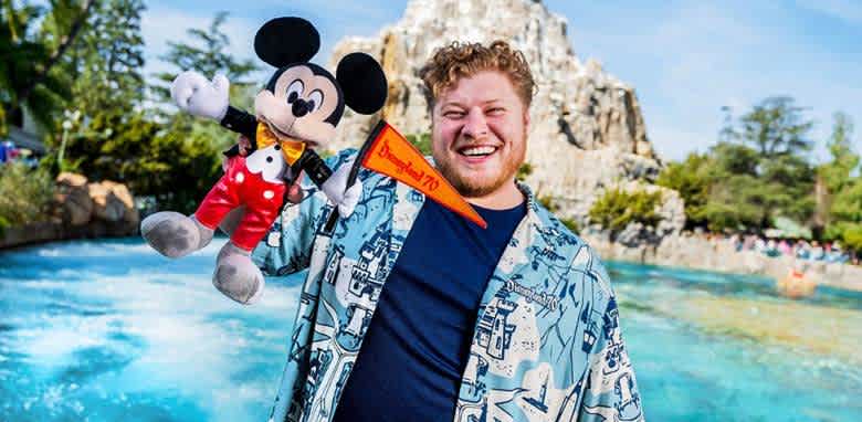 A person stands joyfully by a sparkling blue water feature with a mountain backdrop, holding a stuffed toy resembling a famous mouse character wearing red shorts. They wear a patterned blue shirt and smile widely, conveying a sense of happiness and excitement. Trees and a clear sky complement the lively atmosphere.