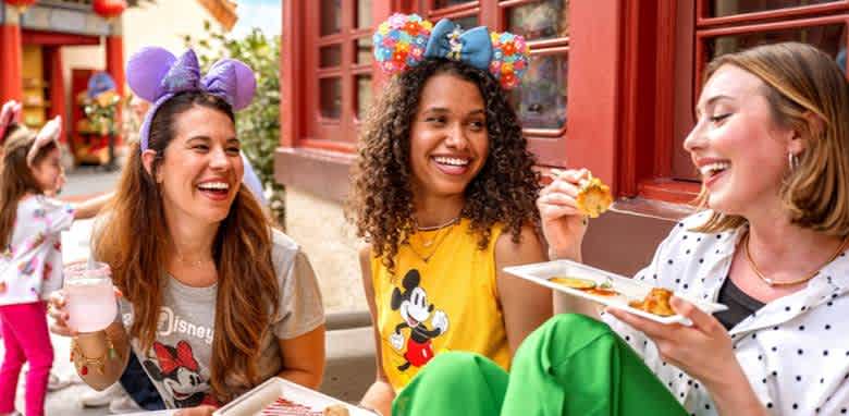 Three friends sit outside enjoying a meal together at a theme park. They wear colorful shirts and mouse ears, smiling and laughing. One holds a drink while another eats from a plate. A child with mouse ears runs in the background. The setting is vibrant and cheerful, suggesting a lively atmosphere filled with fun and excitement.