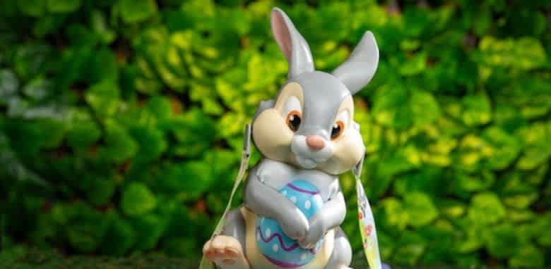 A cute cartoon-style rabbit figurine holding a decorated Easter egg stands against a lush green leafy background. The rabbit has a playful expression with large eyes and soft features. Its fur is a mix of gray and cream colors, and it has large ears. The scene evokes a cheerful, festive atmosphere, ideal for spring celebrations.