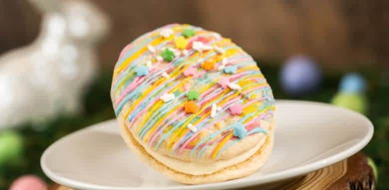 A festive cookie shaped like an Easter egg is displayed on a white plate. The cookie is drizzled with colorful icing and topped with star-shaped sprinkles in various colors. Blurred in the background are pastel-colored eggs and a white ceramic bunny, evoking a cheerful Easter theme.