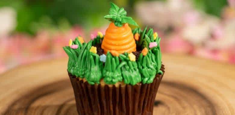 This image shows a cupcake decorated to resemble a garden scene. The cupcake top is adorned with green frosting, mimicking grass, and is accented with small, colorful edible flowers. A carrot-shaped frosting decoration is placed in the center, complete with green leaves on top. The cupcake sits on a wooden surface, and the background is softly blurred with pink, white, and green hues.