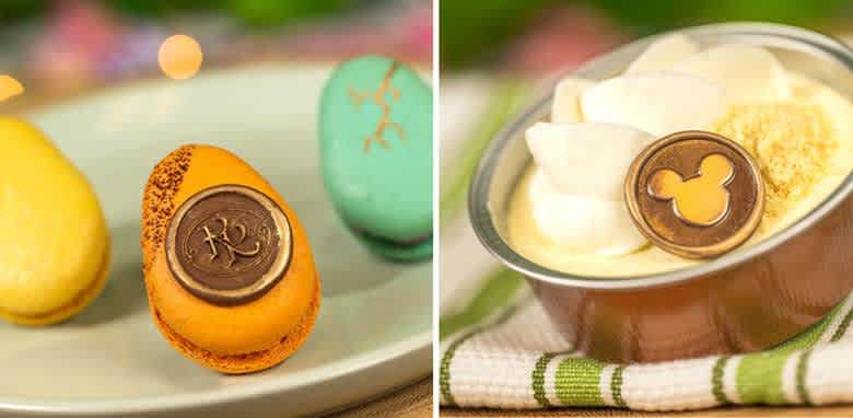 This image shows a plate with colorful macarons. One macaron is orange with a brown chocolate coin embossed with a design. Nearby, a dessert in a silver cup is topped with whipped cream and another chocolate coin featuring a different embossed design. A green and white cloth is partially visible under the cup, against a soft-focus background.
