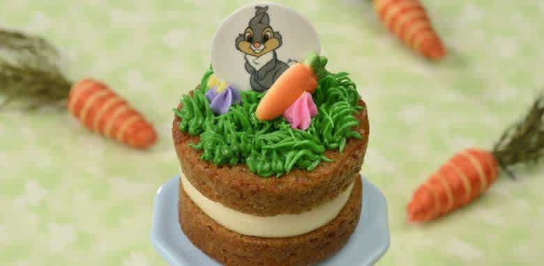 A small two-layer cake topped with green frosting, a fondant carrot, and colorful flowers. There is a round edible image featuring a cartoon character. The background is a soft pattern of carrots, providing a playful and spring-like atmosphere. Perfect for themed celebrations or a fun dessert option.