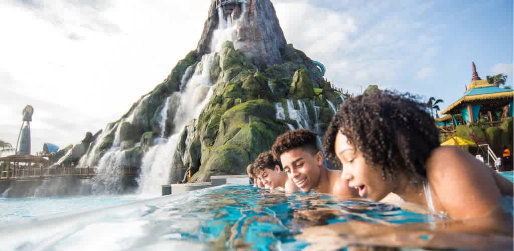 A group of friends enjoys a refreshing moment at a tropical-themed water park in front of a giant volcano waterfall. They happily lean over the edge of a crystal-clear pool, with the lush, green volcano and cascading water creating a thrilling backdrop. Palm trees and vibrant colors enhance the excitement and fun vibe of the scene. Perfect for a memorable adventure.