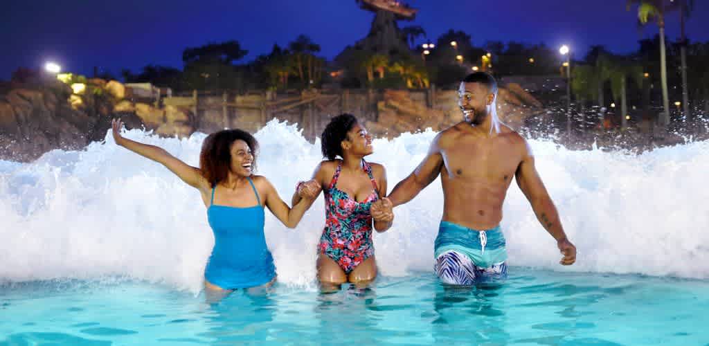 A group of three people enjoys a wave pool at night. They laugh and hold hands while standing in shallow water, with waves crashing around them. The background features a water ride structure and palm trees, all illuminated by ambient lights. The image captures a moment of fun and excitement at a water park.
